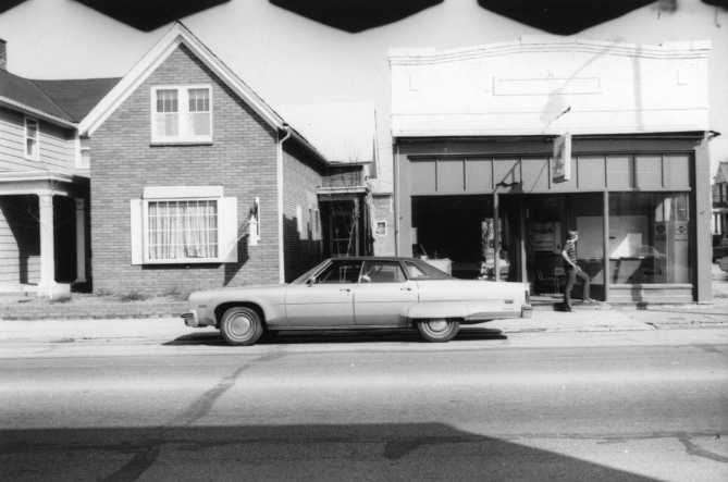 Vintage car parked on a street with buildings and a person in the background. Black and white photo.