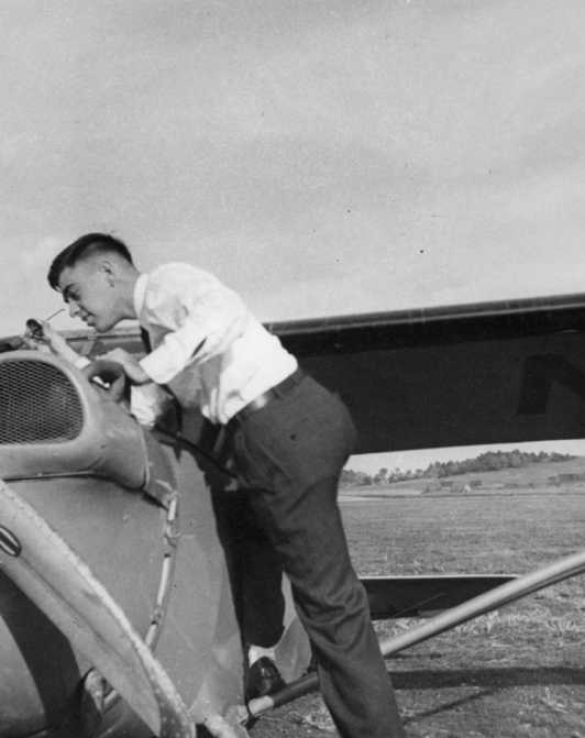 A man works on an old airplane's engine, with open fields in the background.