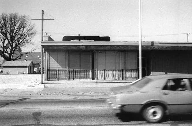 A blurred car passing by a building with barred windows and a vacant signboard, monochrome photo.