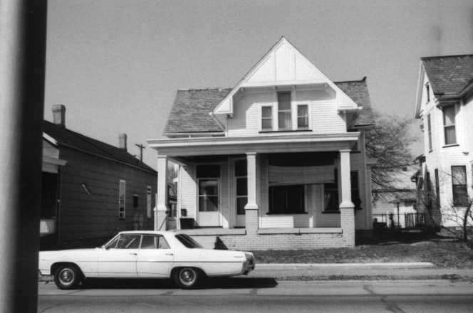 Black and white photo of an old house with a front porch and a vintage car parked out front.