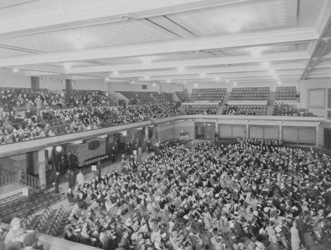 A black and white photo of a crowded auditorium with people seated, facing a stage or speaking area, in a vintage setting.