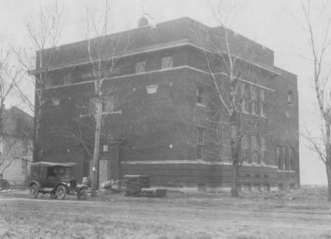 An old black-and-white photo featuring a three-story building, a vintage car, bare trees, and a utility pole.