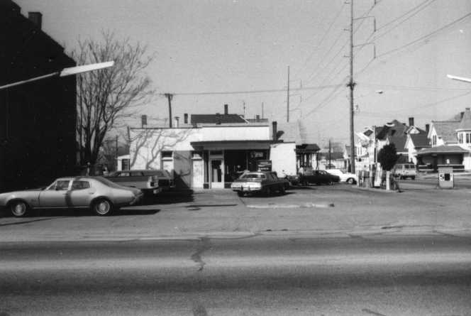 A black and white photo of a vintage gas station with old cars parked nearby and houses in the background.