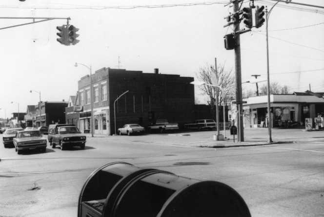 Black and white photo of an urban street corner with vintage cars, traffic lights, buildings, and a mailbox.