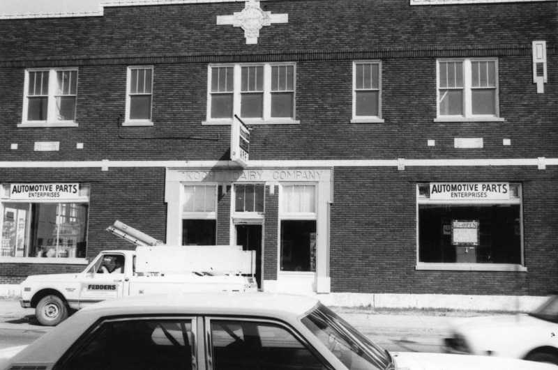 A black and white photo of an old building with signs for Automotive Parts and a utility truck parked in front.