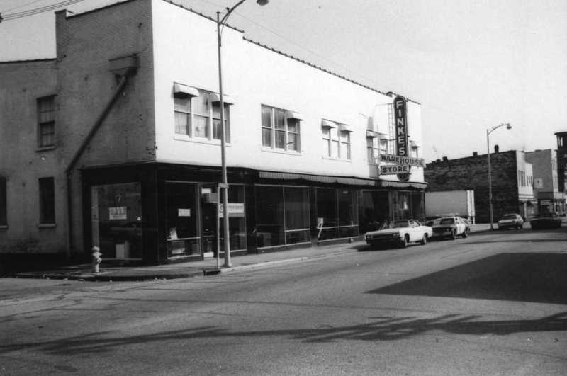 Vintage street view with buildings, signage for "Furniture Warehouse Store," and parked cars.