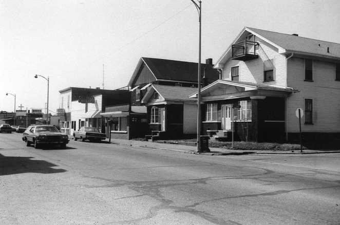 A black and white photo of a quiet street with older houses and a vintage car.