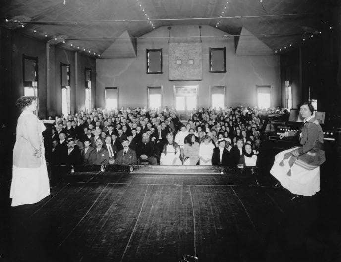 A vintage photo showing a crowded auditorium with two speakers on stage, likely during a public event or assembly.