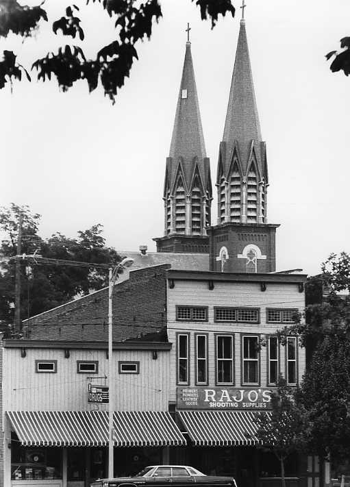 Twin-spired church towering over a storefront with "RAJO'S SHOOTING SUPPLIES" sign, black and white photo. Leafy foreground, vintage car.