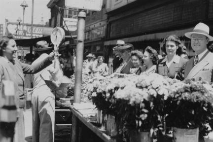 Black and white photo of people on a sunny street, some in hats, looking at flower stalls with an array of blooms.