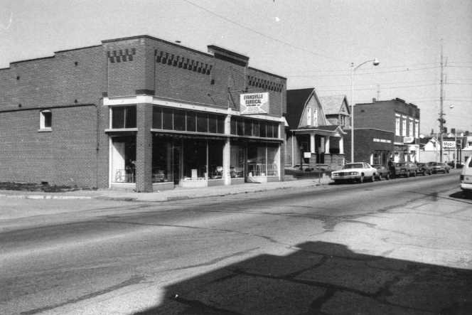 A black and white photo of a vintage street scene with older buildings and cars.