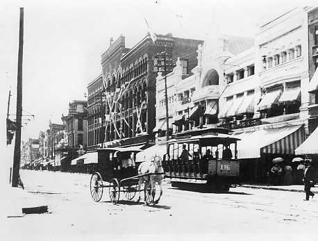 Historic black and white photo of a street scene with a horse-drawn carriage, a streetcar, and vintage buildings.