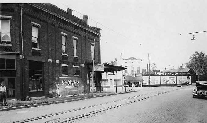 Vintage street scene with brick buildings, a Dr. Pepper sign, tram tracks, and old cars.