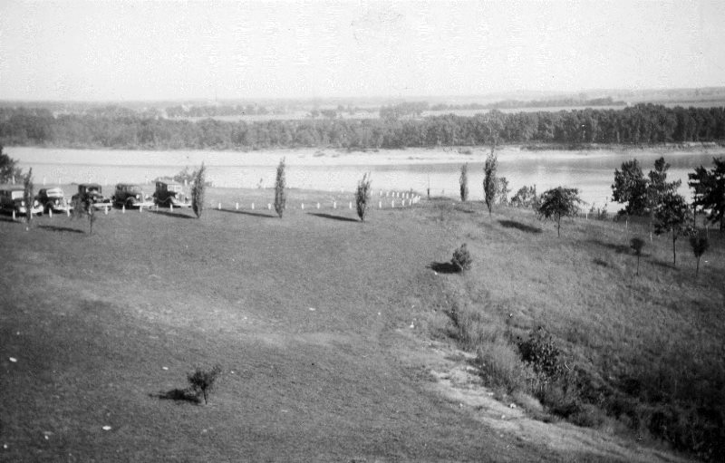 Vintage photo of a riverside landscape with cars parked along a fence.