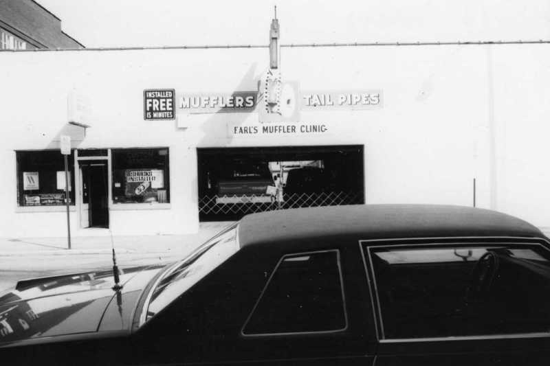 A black and white photo of a car in front of Earl's Muffler Clinic, advertising mufflers and tail pipes.
