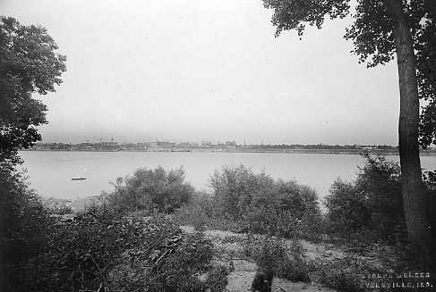 Black and white photo of a river landscape with trees in the foreground and a city skyline in the distance.