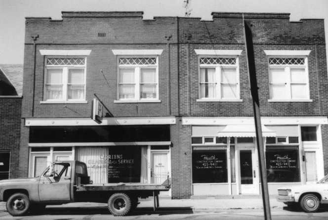 A black and white photo of an older two-story building with a pickup truck parked in front.