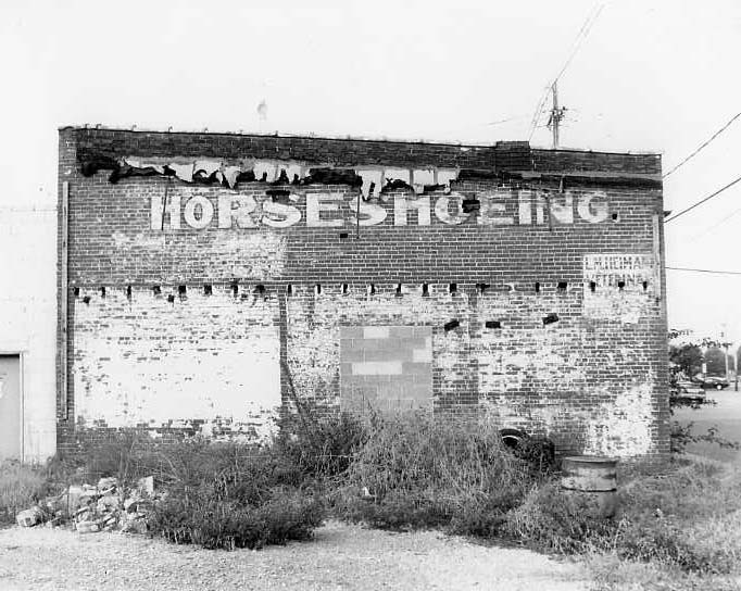 A dilapidated building with faded "HORSE SHOEING" sign, overgrown plants, and a pixelated area on the wall.