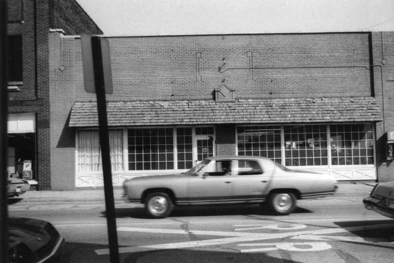Black and white photo of an old building and a vintage car in motion on a city street.