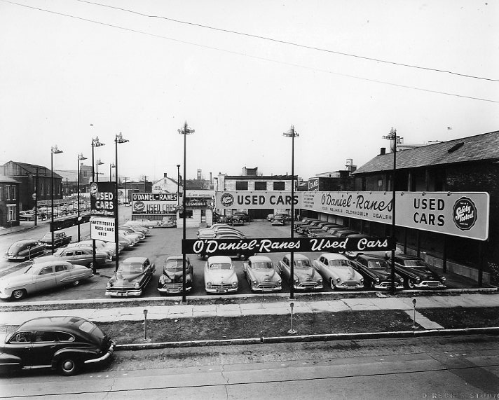 Vintage used car dealership with signs and parked cars.