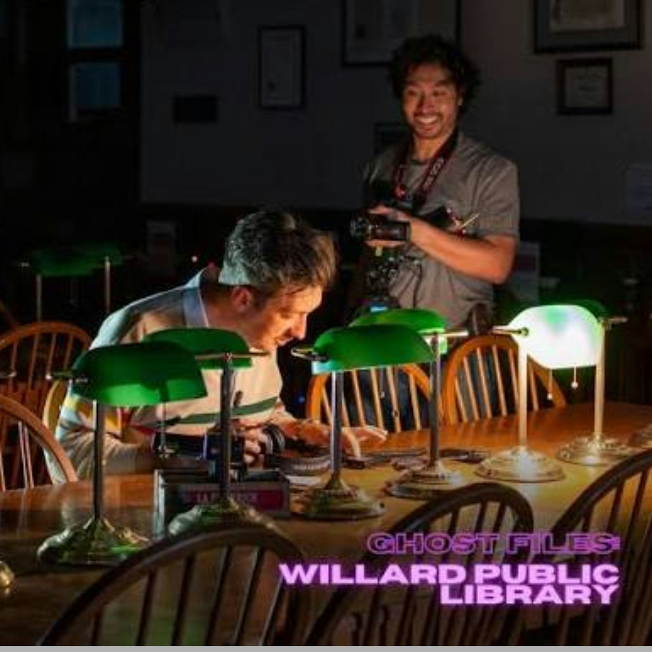 Two men are in a dimly lit library, one examining something at a table with green lamps, while the other takes photos.