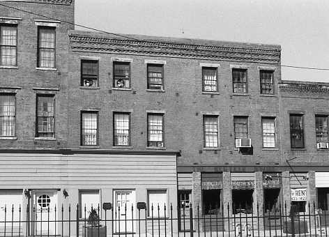 A black and white photo of a three-story urban building with shops on the ground floor, a fenced-off area in front, and a "For Rent" sign visible.