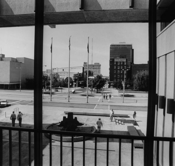 Black and white photo of a cityscape viewed through a frame, with people walking, buildings, and a plaza.