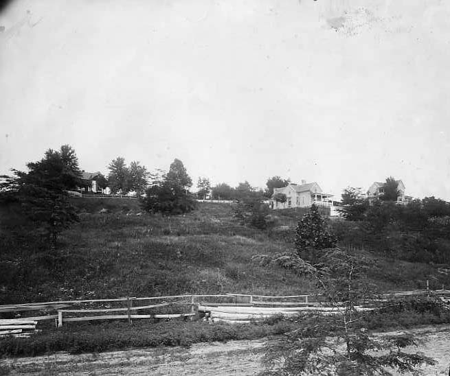 A vintage black and white photo of a rural landscape with houses on a hill and a wooden fence.