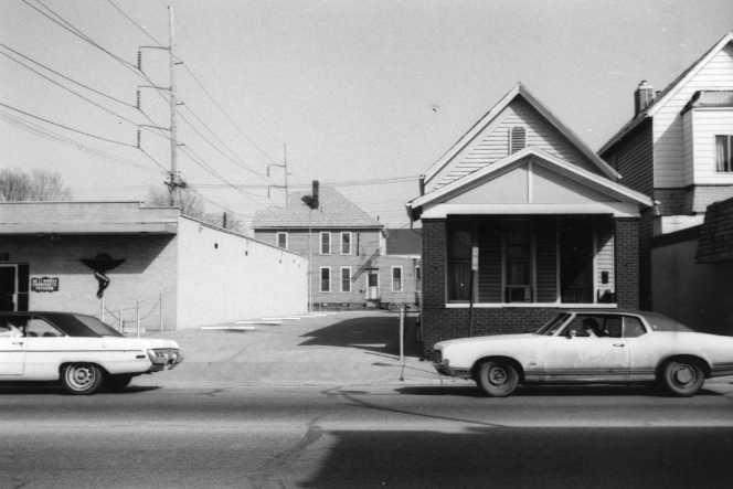 Black and white photo of vintage cars parked by old buildings on a sunny street.