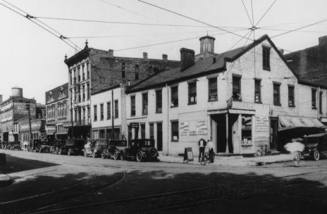 An old black-and-white photo of a street with vintage cars, trolley lines, and historic buildings.
