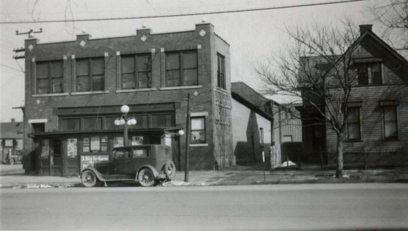 Vintage street scene with an old car parked in front of a two-story building next to a house.