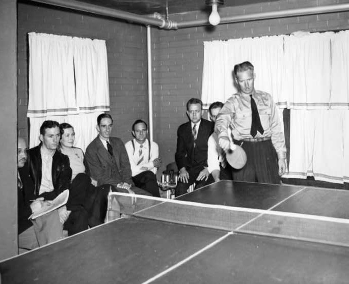 A black-and-white photo of a person playing table tennis, with spectators watching in the background.