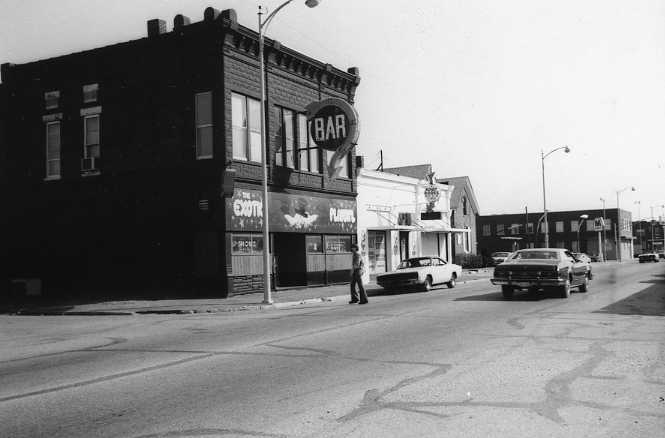 Old black and white photo of a street with cars, a person, and buildings with a bar sign.