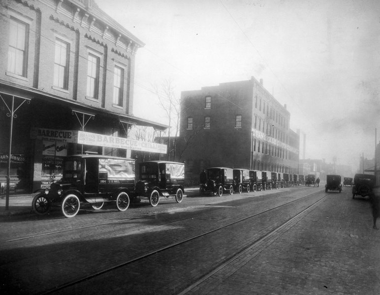 Vintage street lined with early automobiles and brick buildings; streetcar tracks; misty ambiance; "Barbecue" signage.