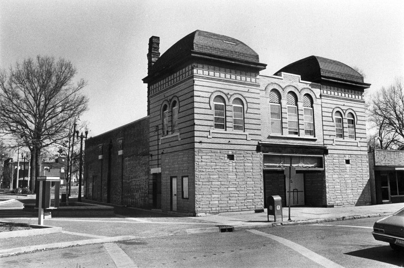 An old building on a street corner with arched windows and a domed tower, next to a leafless tree and a parked car.