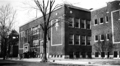 A black-and-white photo of an old school building with people outside.