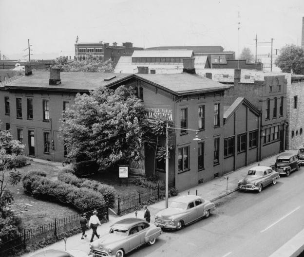 Vintage photo of an urban street with a "Public Museum" sign, old cars parked, and pedestrians walking.