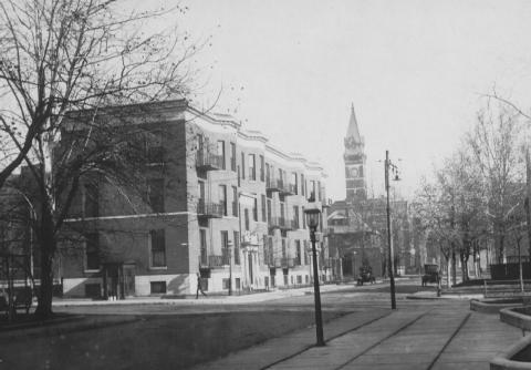 A vintage black-and-white photo of a quiet street with row houses and a church tower in the distance.