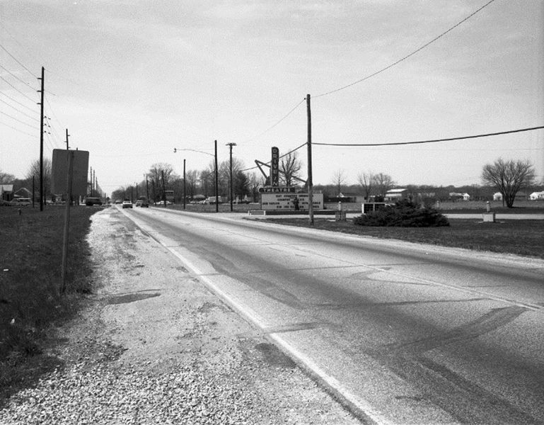 Black and white photo of an empty road with power lines, a sign for a motel and some trees.