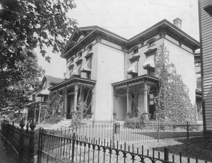 Historic two-story house with large windows, columns, and ivy, surrounded by a wrought iron fence.