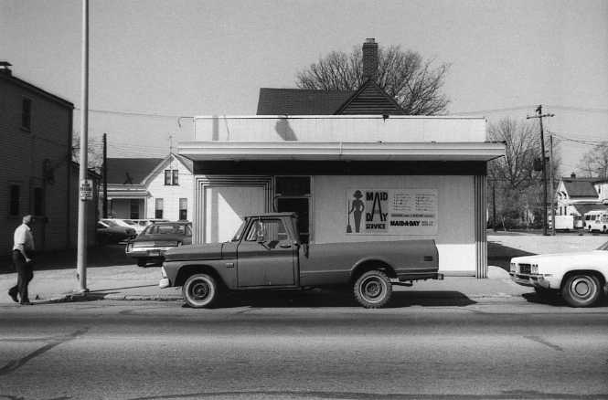 Black and white photo shows a street scene with a small building, two older vehicles and a pedestrian.