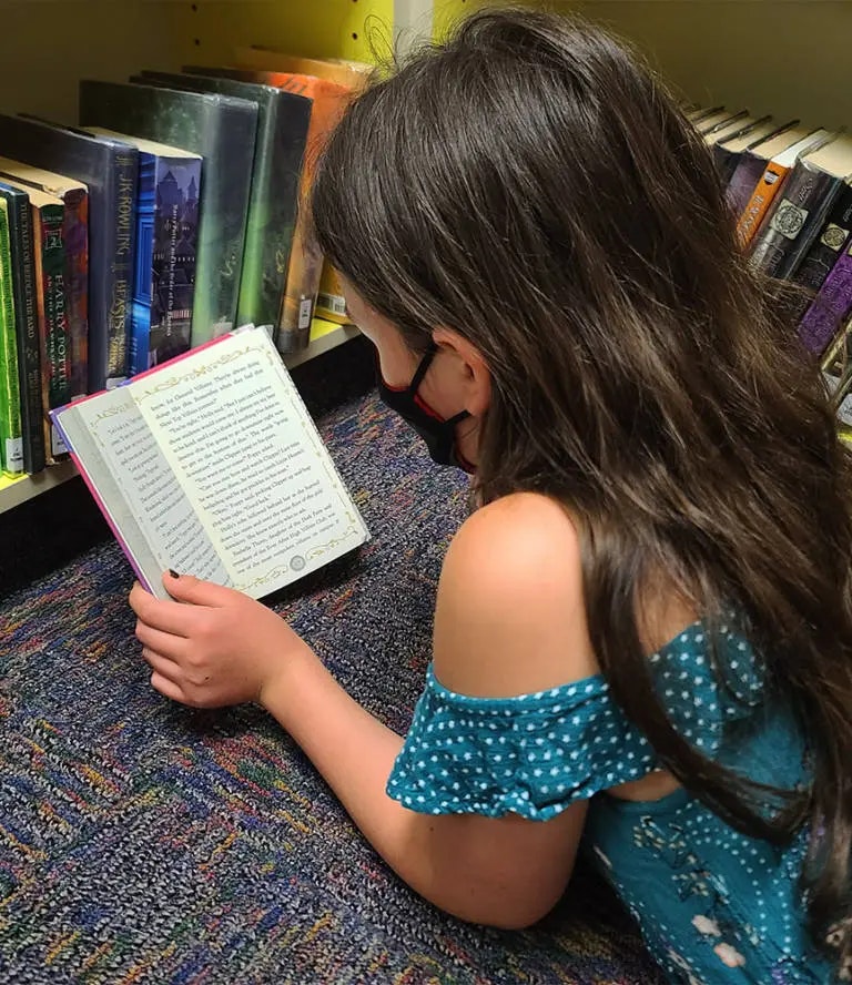 A girl is reading a book in a library aisle, with shelves of books visible.