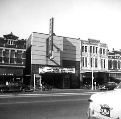 A vintage black and white photo of a classic movie theater facade on a city street with cars and a pedestrian.