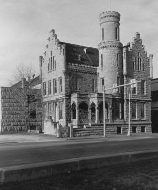 Historic building resembling a small castle with a turret, arched windows, and stone walls, situated beside a street.