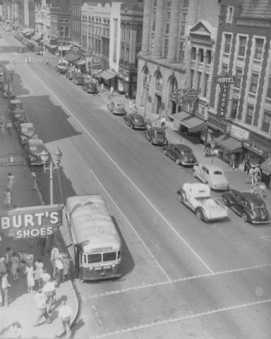 Vintage street scene with old cars, bus, signs for businesses like "BURT'S SHOES", and buildings. Likely mid-20th century.
