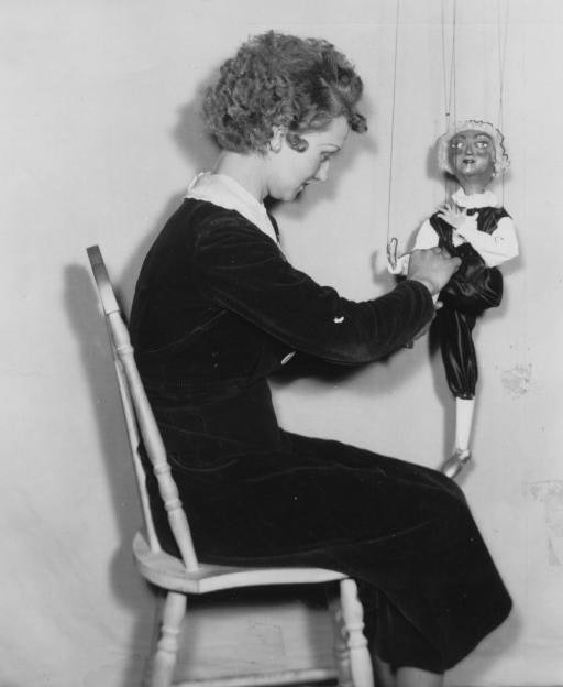 A woman is sitting, manipulating a marionette puppet against a plain background.