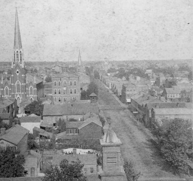 Vintage photo of a town with buildings, church spire, and tree-lined street.