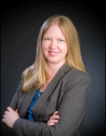 A professional headshot of a woman with long hair, wearing a suit and blue blouse, arms crossed, on a black background.