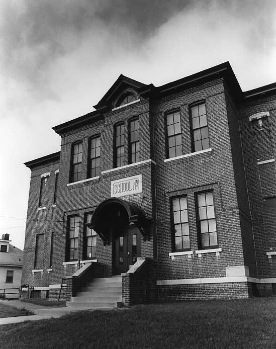 Black and white photo of an old brick school building with stairs and arched entryway under a cloudy sky.