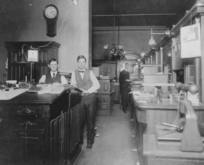 Vintage photo of men in an old-fashioned office with wooden desks, clock on wall, and gas lamps.